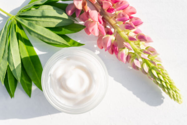 Open jar of white moisturizer cream in a clear container placed on a white surface, accompanied by a lush green leaf and a pink flowering lupine stem, symbolizing natural skincare ingredients and botanical beauty enhancements.